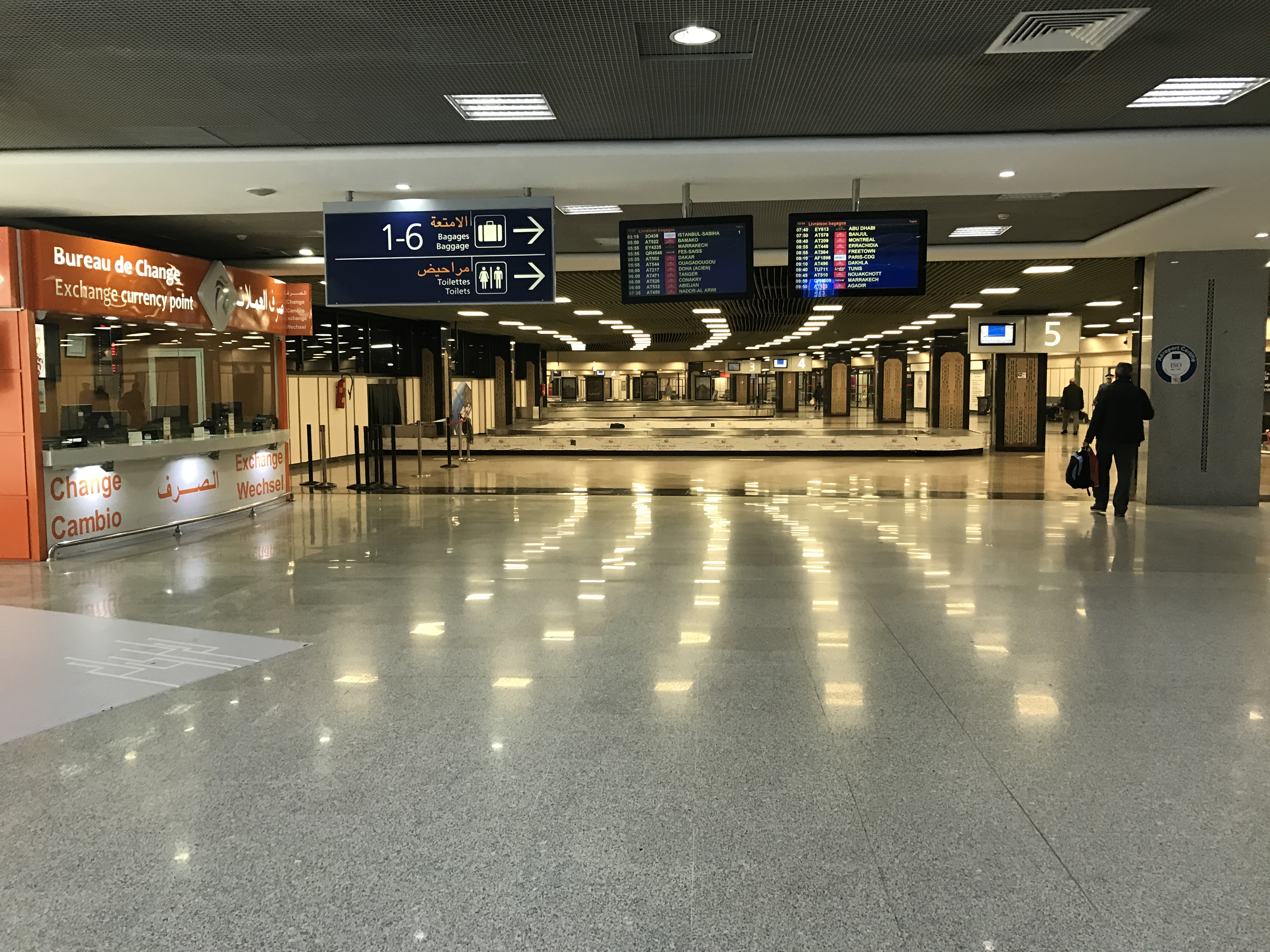 An empty baggage carousel in an airport arrivals hall with a few travelers waiting nearby.