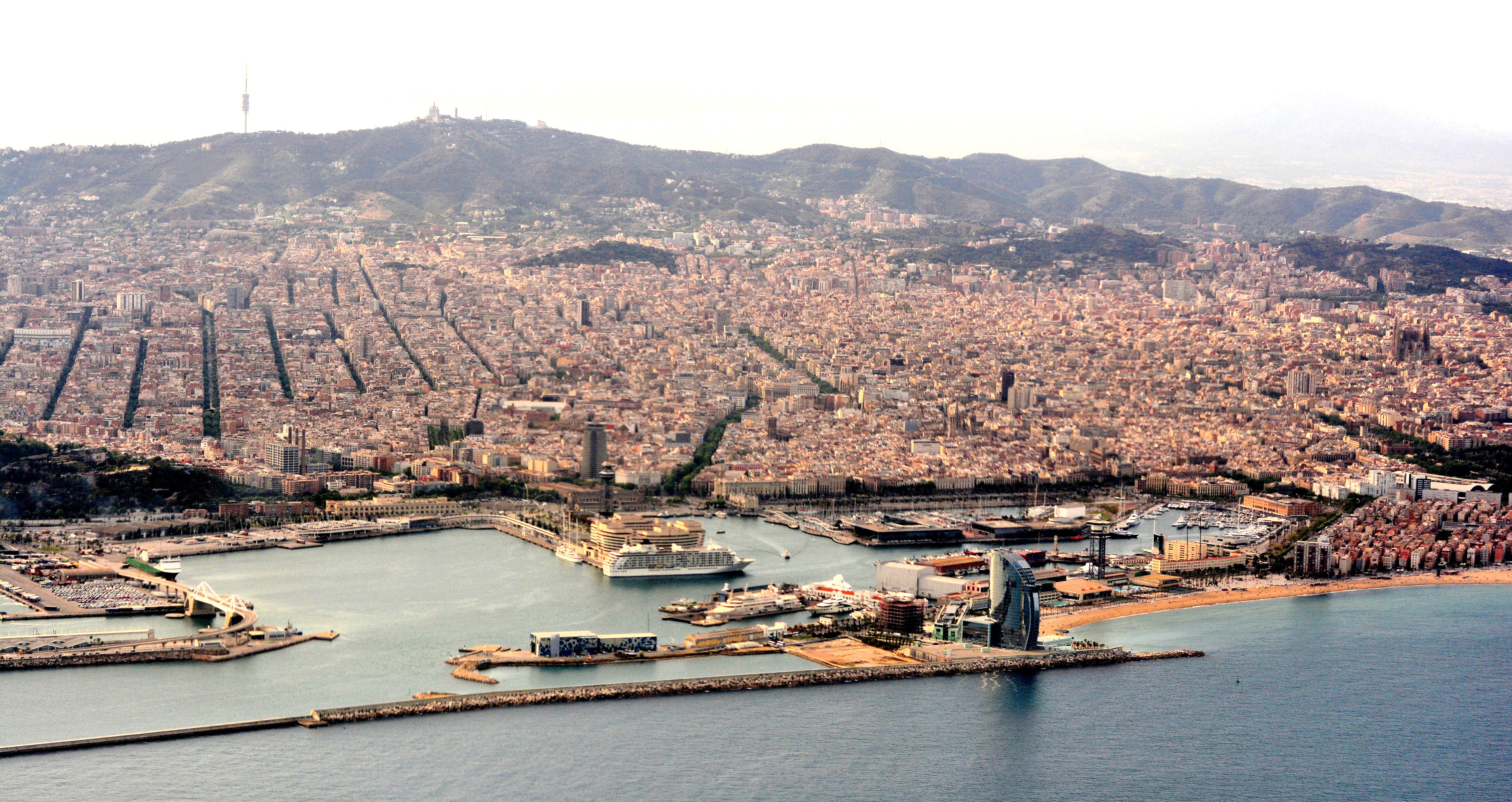 A large cruise ship docked at a sunny Mediterranean port with the city skyline behind it.