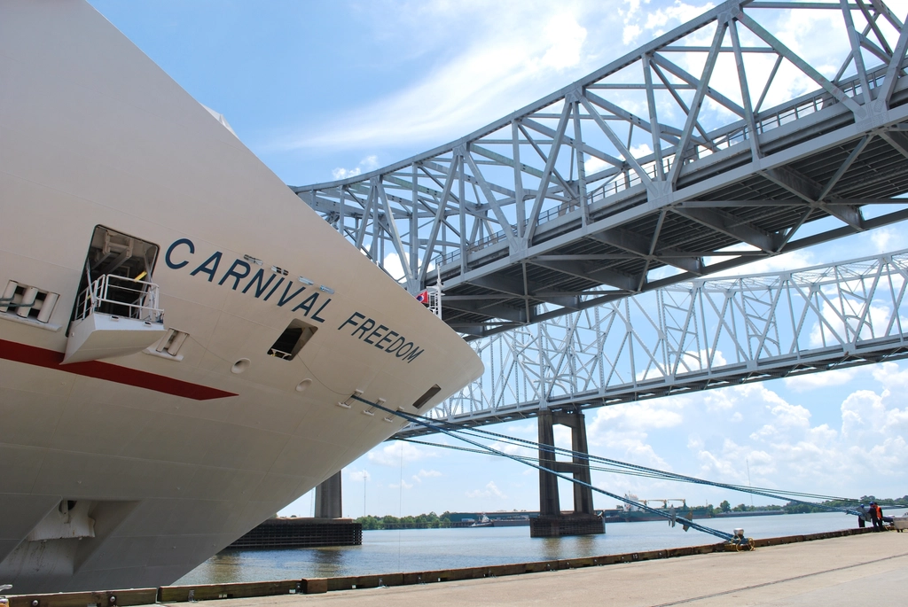 A large cruise ship docked at a port terminal while a passenger jet passes overhead.