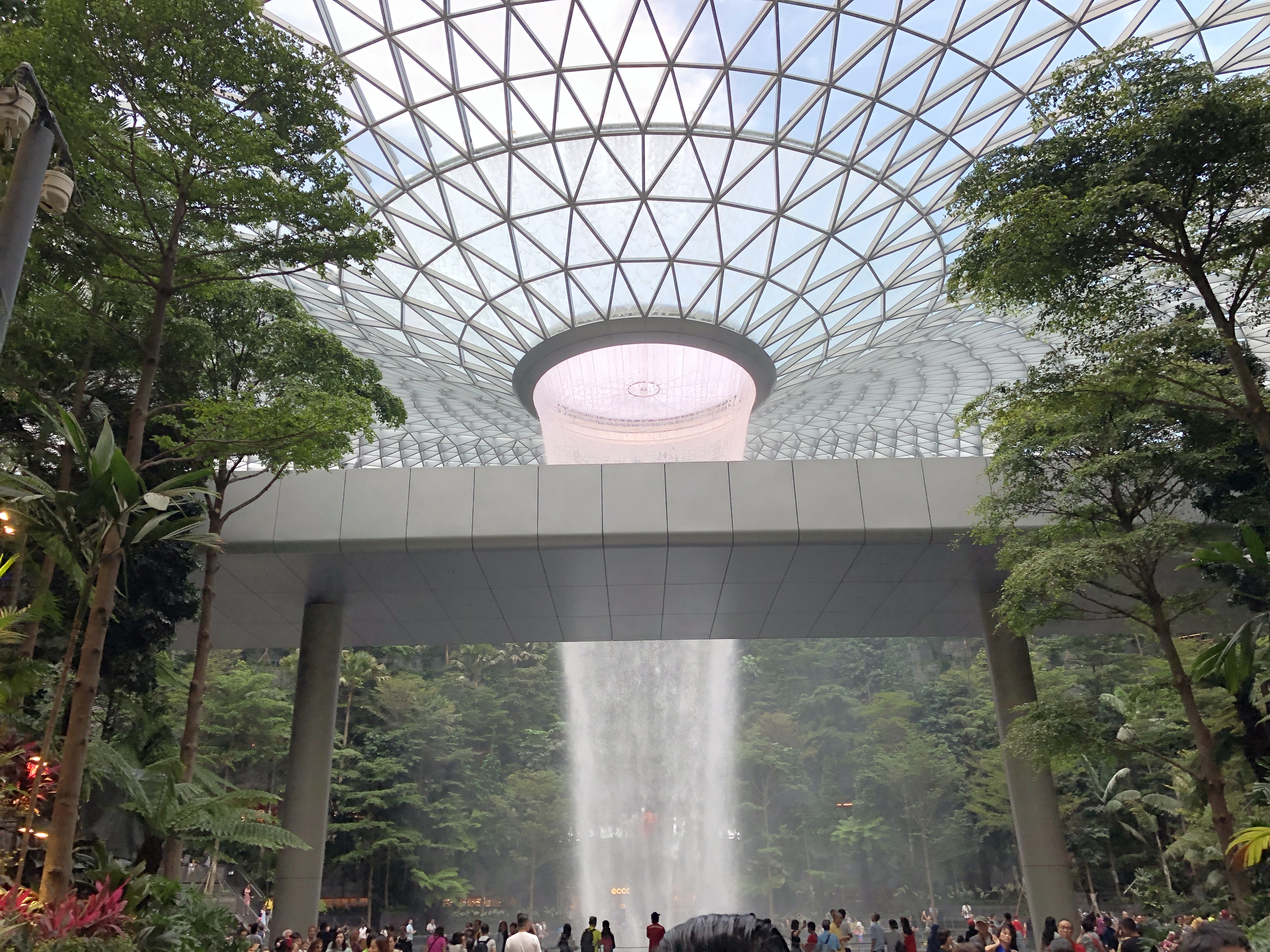The Rain Vortex indoor waterfall pouring through the glass dome of Jewel Changi Airport, surrounded by terraced indoor forest.